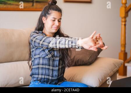 Sorridente giovane freelance in abito casual seduto sul divano di fronte al computer portatile e che si allunga mentre si riposa durante il lavoro su progetto a casa Foto Stock