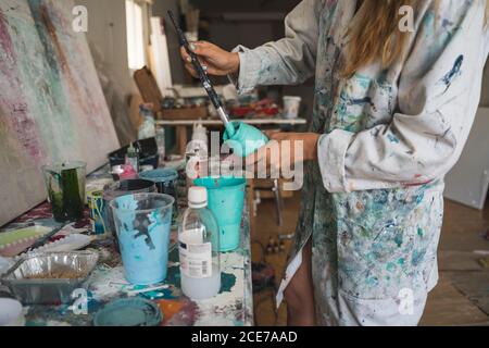 Vista laterale ad alto angolo del raccolto giovane artista femminile in piedi al contatore con vari strumenti e la preparazione dei colori per il disegno mentre si lavora in un'officina creativa Foto Stock