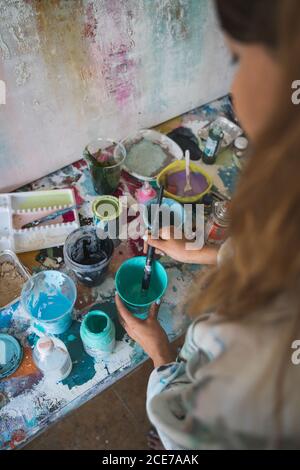 Vista laterale ad alto angolo del raccolto giovane artista femminile in piedi al contatore con vari strumenti e la preparazione dei colori per il disegno mentre si lavora in un'officina creativa Foto Stock