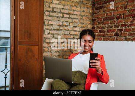 Felice elegante African American Woman messaggio di controllo sul telefono cellulare mentre si siede con il portatile in poltrona contro muro di mattoni in appartamento loft Foto Stock