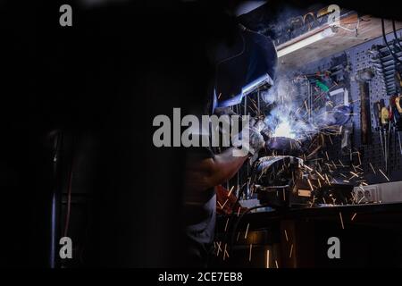 Vista laterale di un operaio maschile anonimo in piedi casco protettivo al banco di lavoro e fissaggio dei dettagli metallici con l'utensile di saldatura Foto Stock