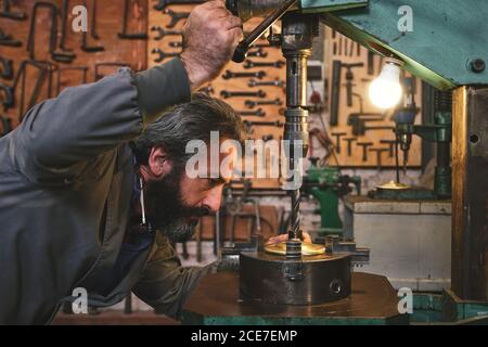 Vista laterale di un artigiano serio concentrato di mezza età in uniforme utilizzo di una macchina da incisione mentre si lavora con il metallo in studio Foto Stock