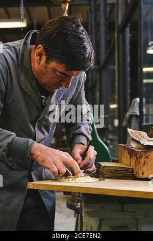 Orafo etnico di media età concentrato in uniforme lavorando con oro su tavola di legno usando bastone di metallo mentre si inclina in avanti dentro studio Foto Stock