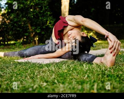 Giovane slim femminile in abbigliamento sportivo che pratica Parivrtta Upavistha Konasana yoga asana su erba verde nel parco estivo Foto Stock