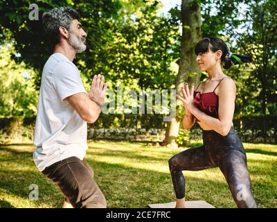 Vista laterale del maschio adulto bearded e della donna slim in abbigliamento sportivo in piedi faccia a faccia in posa Dea mentre pratica yoga insieme nel parco nel giorno d'estate Foto Stock