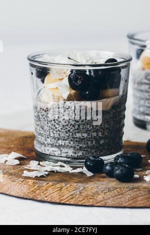 Vasetti di vetro con deliziosi budini chia fatti di fresco gustoso latte di mandorle e semi di cia con miele condito con more e la banana affettata servita su tavola di legno su sfondo bianco Foto Stock