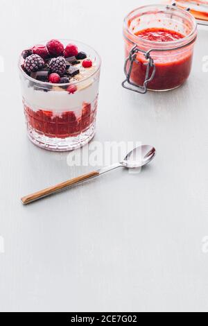 Bicchiere di gelato con marmellata di fragole, frutti di bosco surgelati e patatine di cioccolato posto su un tavolo bianco di legno Foto Stock