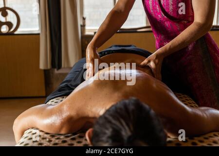 Crop anonimo master eseguire massaggio ayurvedico con olio aromatico ON retro di cliente rilassato in un salone moderno Foto Stock