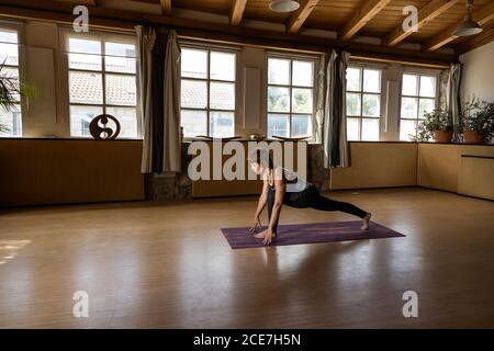 Vista laterale della donna dai capelli rossi che pratica yoga sul suo tappeto nel suo studio di legno e pietra, vista laterale Foto Stock