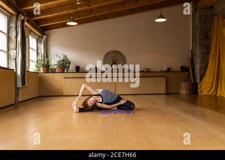 Femmina in abbigliamento sportivo seduto in rotazione angolo seduto mentre si pratica Shakti yoga da solo in studio luminoso con interni in legno resort tropicale Foto Stock