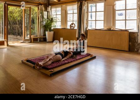 Allenatore femminile aiutare flessibile donna braccio stretching in Eka Pada Pavanamuktasana mentre fare yoga in studio Foto Stock