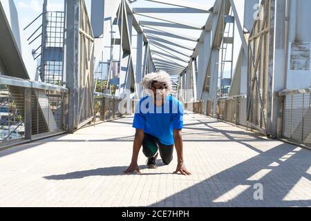 Atleta nero con capelli biondi che prendono la posizione iniziale del crouch prima srotolando su un moderno ponte in una giornata di sole in città Foto Stock