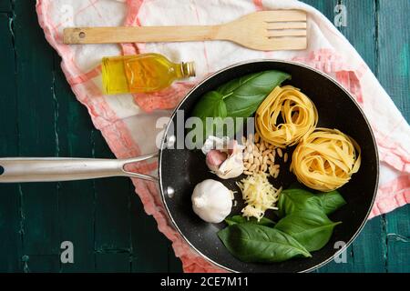 Vista dall'alto della penna per friggere con panini alla pasta non cotti nelle vicinanze foglie di basilico fresco e teste di aglio con pinoli e bottiglia di olio d'oliva vicino a spatola di legno su asciugamano Foto Stock