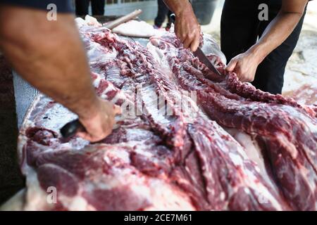 Angolo alto di macellai anonimi di raccolto che tagliano la carne di maiale sopra tavola in legno durante la produzione di prodotti tradizionali in azienda Foto Stock