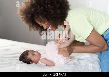 Vista laterale di allegra Afro American Woman divertirsi con adorabile neonato sdraiato su un morbido letto Foto Stock