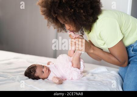 Vista laterale di allegra Afro American Woman divertirsi con adorabile neonato sdraiato su un morbido letto Foto Stock
