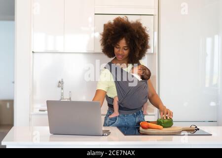 Madre afroamericana che trasporta il bambino addormentato nell'imbragatura e nel taglio verdure fresche in cucina mentre si prepara gustosa ricetta di osservazione del pranzo su un computer portatile Foto Stock