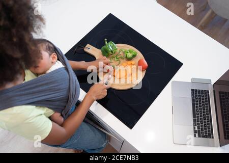 Dall'alto di madre afroamericana che trasporta il bambino che dorme dentro imbragare e tagliare verdure fresche in cucina mentre si prepara gustoso pranzo Foto Stock