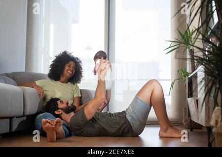 Soddisfare la madre multirazziale e padre che gioca con il bambino adorabile mentre riposante su un piano in un appartamento moderno Foto Stock