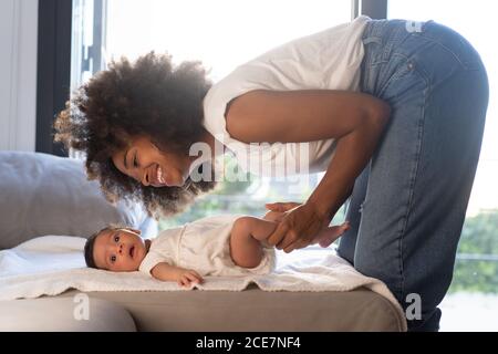 Vista laterale di una donna afro-americana giochissima che gioca con un bambino carino sul divano di casa Foto Stock