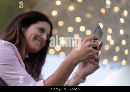 Vista laterale basso angolo del contenuto femmina che scatta foto sopra fotocamera del telefono cellulare su sfondo sfocato di ghiaino incandescente Foto Stock