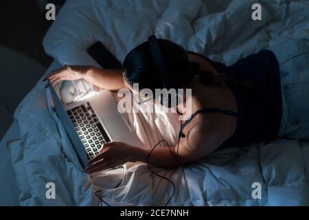 Vista dall'alto sul retro di una femmina rilassata nella parte superiore del serbatoio sdraiato su una coperta sul letto mentre si ascoltano le canzoni laptop in cuffia in serata Foto Stock