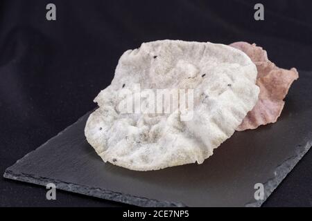 Cracker sottili di riso fritto croccante, croccanti e di sapore semplice, di colore bianco e viola, a base di riso, sesamo, sale su una piastra di pietra nera su sfondo nero Foto Stock