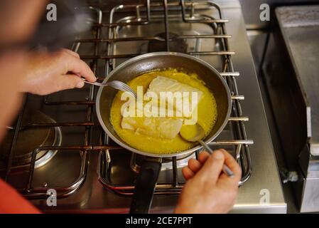 Dall'alto di raccolto unriconoscable chef passando filetti di pesce dentro sciogliere il burro e l'olio d'oliva in padella utilizzando la forchetta e cucchiaio mentre si prepara il cibo a casa Foto Stock