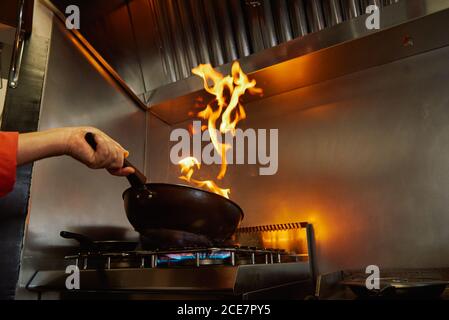 Da sotto, uno chef non riconoscibile e tagliato in padella di frittura uniforme mentre gli ingredienti si infiammano in padella a fuoco lento in cucina ristorante in serata e guardando fuoco luminoso Foto Stock