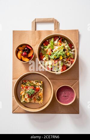 Vista dall'alto dell'insalata di verdure con vari ingredienti nutrienti in recipiente di cartone posto su sacchetto di carta vicino al riso con Molí de Pals riso vegetale e crema di barbabietole e frutta ciotola Foto Stock