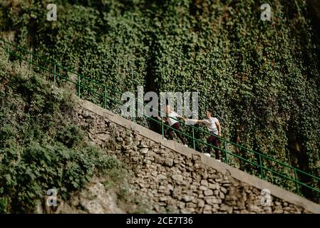 Vista laterale basso angolo di giovane uomo seguendo allegra ragazza mentre salite le scale vicino ai cespugli verdi durante il viaggio emozionante in natura Foto Stock