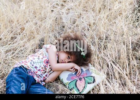 Adorabile ragazza mulatto calmo in abito trendy sdraiato sulla plaid sul campo di grano il giorno d'estate e guardando la macchina fotografica Foto Stock