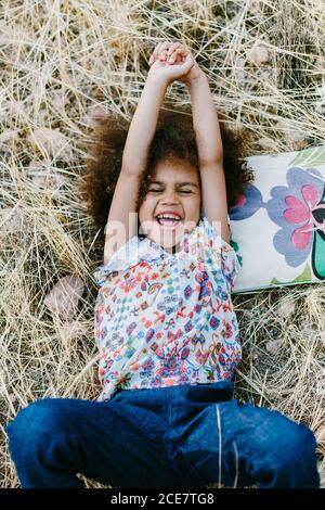 Adorabile ragazza mulatto calmo in abito trendy sdraiato sulla plaid sul campo di grano il giorno d'estate e guardando la macchina fotografica Foto Stock