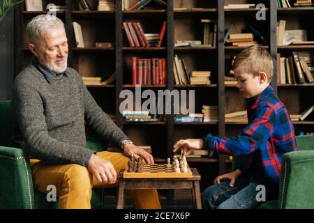 Sorridendo felice anziano nonno e nipote stanno giocando a scacchi seduto alla scrivania Foto Stock