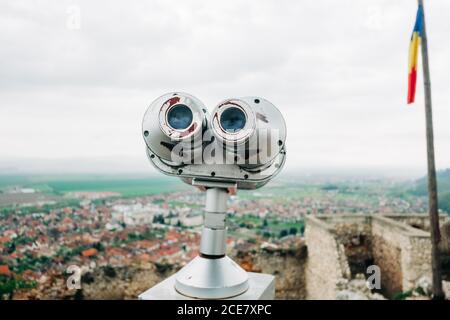 Binocolo panoramico stazionario situato sulla piattaforma di osservazione vicino alla bandiera rumena Contro l'accogliente città vecchia in Transilvania in giorno nuvoloso Foto Stock