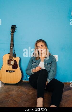 Giovane musicista femminile pensieroso seduto accanto alla chitarra mentre si siede pavimento contro parete blu che guarda via Foto Stock