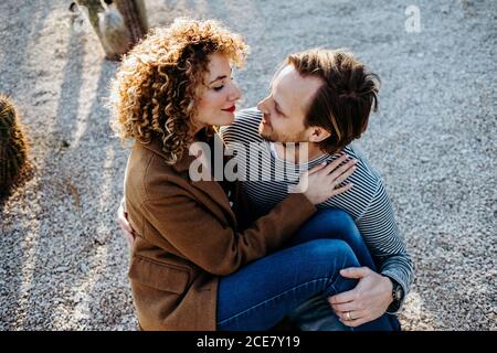 Vista dall'alto dell'allegro uomo e della donna sorridenti e che si abbracciano seduti sul pavimento mentre si passa la giornata estiva sullo sfondo sfocato dei cactus nel parco di Barcellona, Spagna Foto Stock