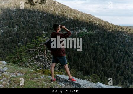 Vista posteriore di turista maschile irriconoscibile con zaino in piedi pendenza della montagna e vista delle verdi colline mentre osservano Paesaggio con binocoli in Navacerrada in Spagna Foto Stock