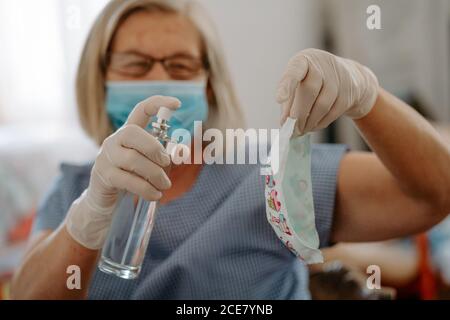 Master senior femminile in maschera protettiva e guanti in lattice disinfezione maschera in tessuto fatta a mano con igienizzatore mentre si producono maschere per coronavirus prevenzione sul luogo di lavoro Foto Stock