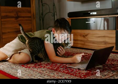 Allegro adorabile bambino coccolando gatto e divertirsi mentre si siede sul pavimento e guardando i video su un computer portatile Foto Stock