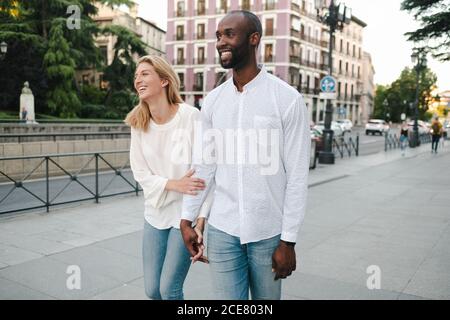 Ottimista sorridente coppia multirazziale che indossa jeans e camicie bianche a piedi Lungo la strada moderna di Madrid e tenendo le mani teneramente dentro giorno Foto Stock