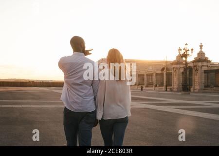 Back view coppia eterogenea anonima in abiti casual che camminano insieme lungo il marciapiede asfaltato sulla strada durante il tramonto Foto Stock