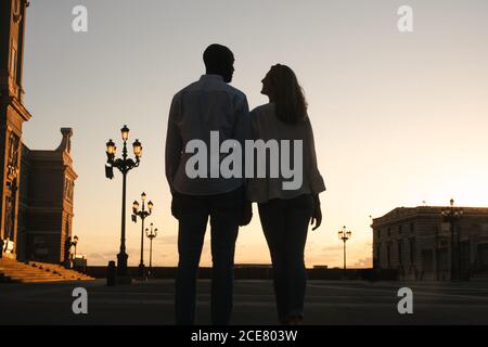 Back view coppia eterogenea anonima in abiti casual che camminano insieme lungo il marciapiede asfaltato sulla strada durante il tramonto Foto Stock