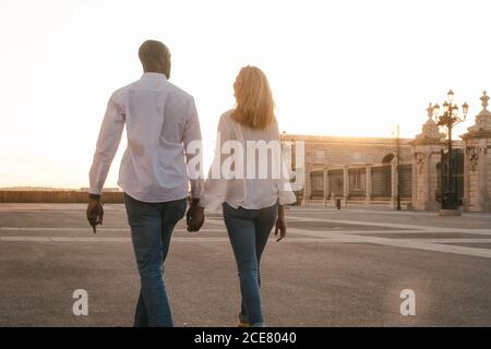 Back view coppia eterogenea anonima in abiti casual che camminano insieme lungo il marciapiede asfaltato sulla strada durante il tramonto Foto Stock