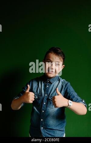 Allieva allegro ragazzo di grado che indossa una camicia in denim mostrando le mani con i pollici verso l'alto gesto e guardando la fotocamera mentre sorridi e. in piedi su sfondo verde scuro Foto Stock