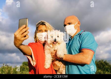 Contenuto Coppia adulta in maschere di faccia che indossa abiti estivi casual In piedi con adorabile cane bianco Bichon Frise sulle mani e. prendere selfie sul telefono cellulare mentre si passa la sera in natura Foto Stock