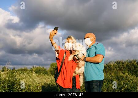 Contenuto Coppia adulta in maschere di faccia che indossa abiti estivi casual In piedi con adorabile cane bianco Bichon Frise sulle mani e. prendere selfie sul telefono cellulare mentre si passa la sera in natura Foto Stock