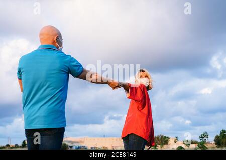 Romantica coppia di mezza età in respiratori e vestiti casual tenuta mani e filatura mentre camminando insieme in campagna durante l'estate sera e guardarsi l'un l'altro con amore Foto Stock