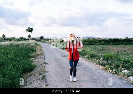 Donna positiva in abiti casual indossando maschera protettiva in piedi Con divertente cane bianco Bichon Frise sulle spalle contro sfocato campagna estiva asfalto strada paesaggio durante l'epidemia di coronavirus Foto Stock