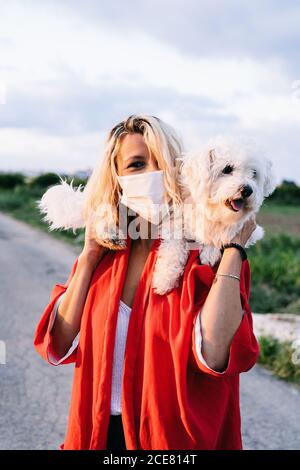 Donna positiva in abiti casual indossando maschera protettiva in piedi Con divertente cane bianco Bichon Frise sulle spalle contro sfocato campagna estiva asfalto strada paesaggio durante l'epidemia di coronavirus Foto Stock